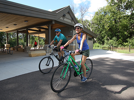 Two people riding bikes in front of a picnic shelter.