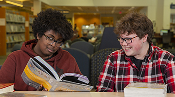 Two teens at a table reading a book and smiling.