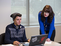 Teen boy looking at a computer screen with a tutor looking over his shoulder. .