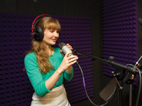 Woman standing at a microphone wearing headphones.