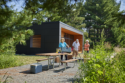 Three people setting up at a picnic table in front of a camper cabin.